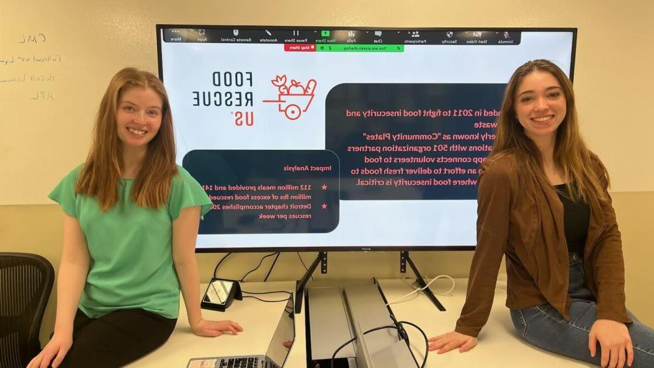 韦德体育app官网 学生s, Sarah Nyquist and Rachel Whalen, 坐在一个大显示器前，上面显示着美国食品救援组织的信息.